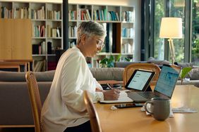 older woman takes notes while working on laptop and tablet with charts on screen