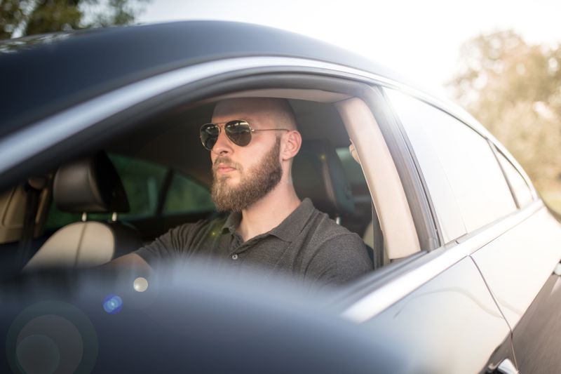 A bearded driver wears sunglasses in a black car