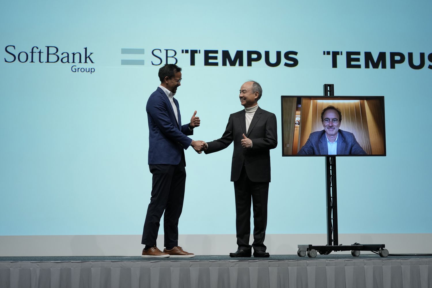 Masayoshi Son, chairman and chief executive officer of SoftBank Group Corp., center, shakes hands with Ryan Fukushima, left, chief operating officer of Tempus AI Inc., as Eric Lefkofsky, chief executive officer of Tempus AI Inc., on a monitor during a news conference in Tokyo, Japan.