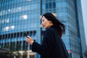 A young professional woman checks her smartphone while standing in front of a glass-front skyscraper.