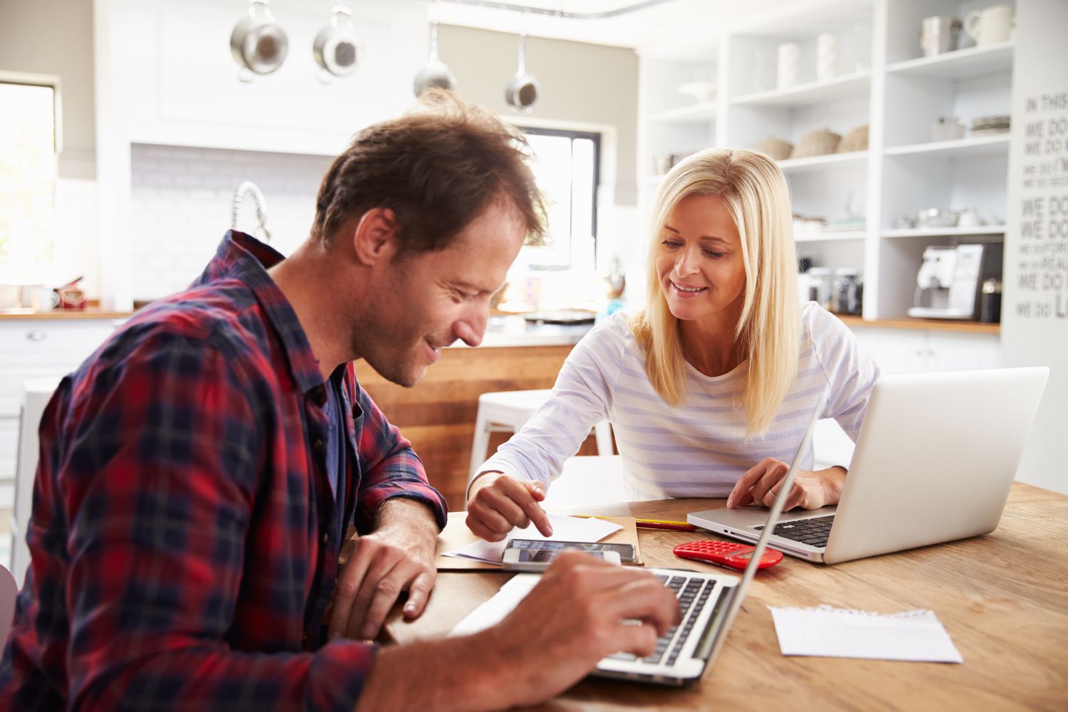 Couple in their 30s sitting in their kitchen and looking at laptops and a calculator.