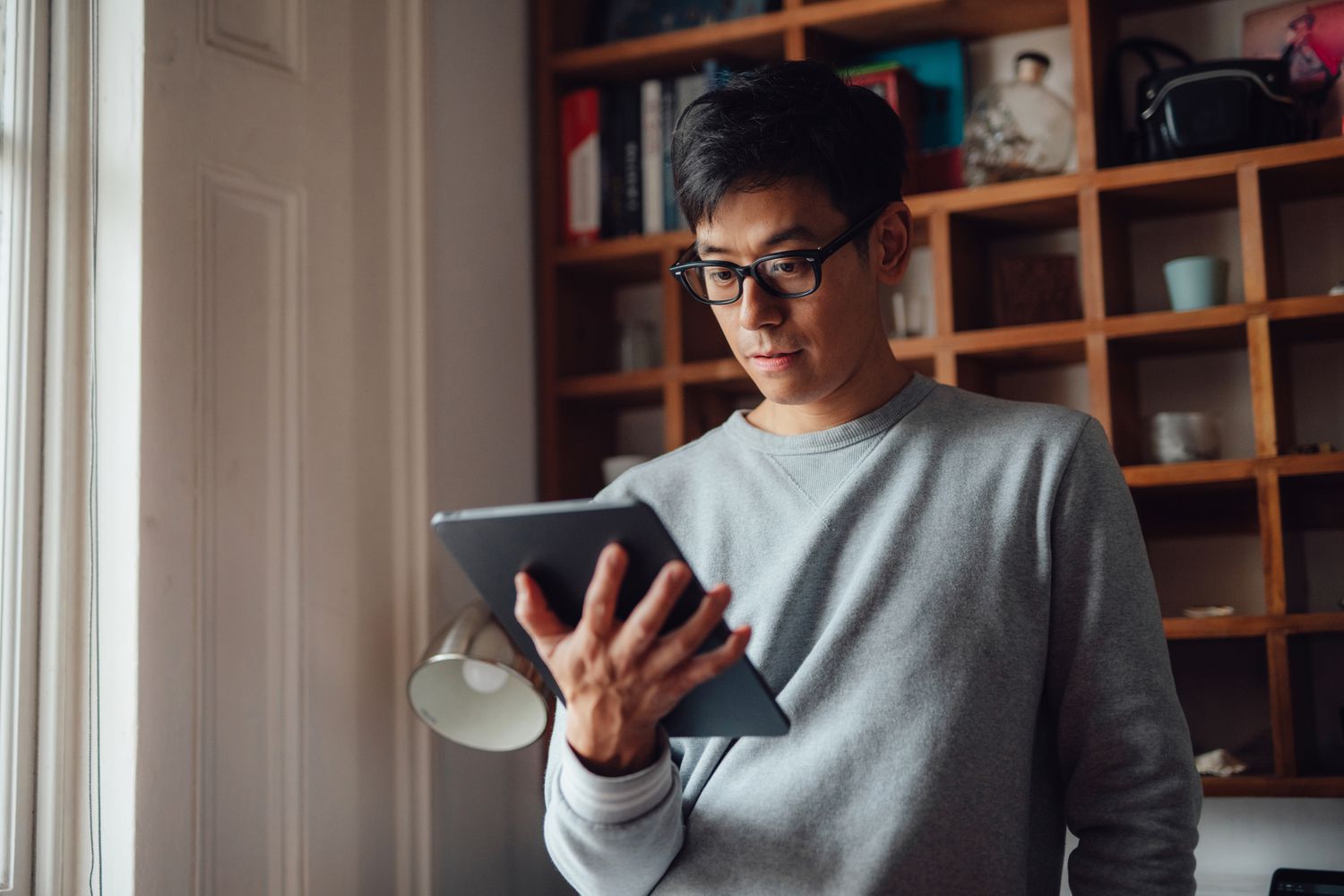 Low angle view of young professional Asian man with glasses, using digital tablet by the window at home. 