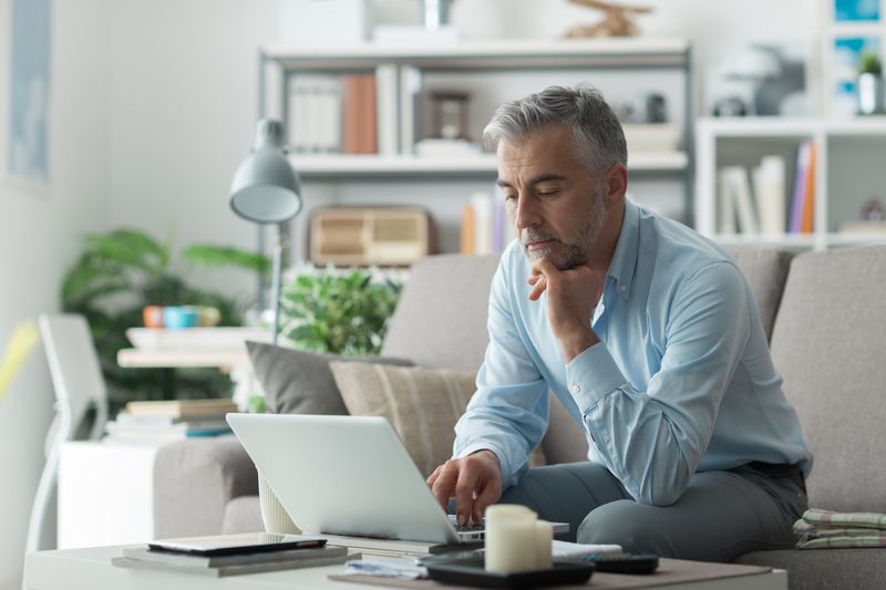Man in his 50s sitting on his sofa and looking intently at financial information on his laptop