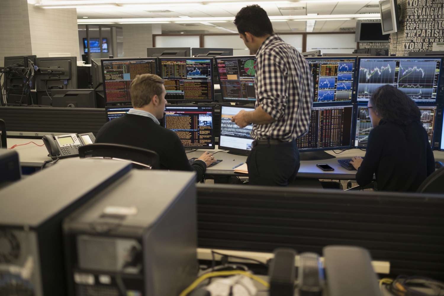 A team of three traders discuss strategies and trade execution in front of computer monitors displaying different information. 