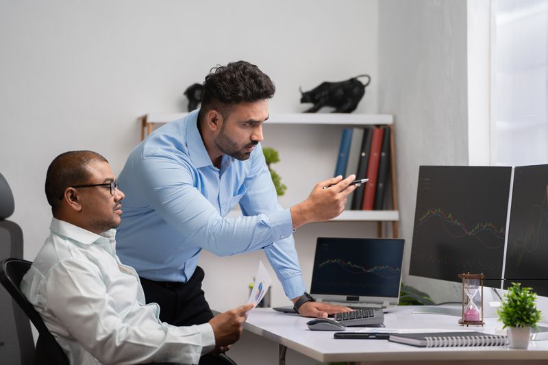 A pair of traders use desktop computers and discuss forming a short straddle strategy for a security they follow.