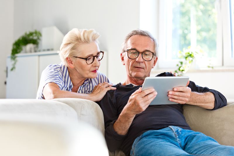 Middle-aged man reclining on a white couch holding a tablet. His wife squatting behind the couch is looking over his shoulder at the tablet. 