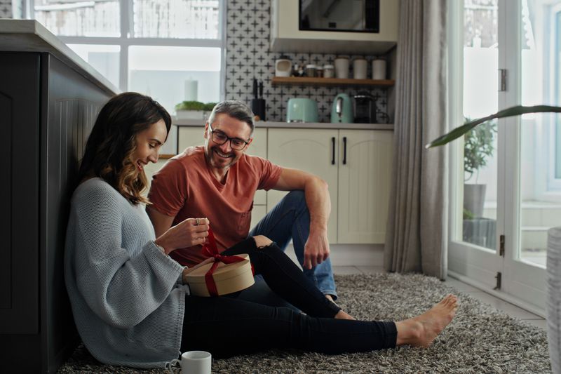 Woman opening a gift while man watches her, smiling, both sitting on the kitchen floor