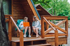 Two women sitting on chairs outside of a cabin, with one holding a laptop.