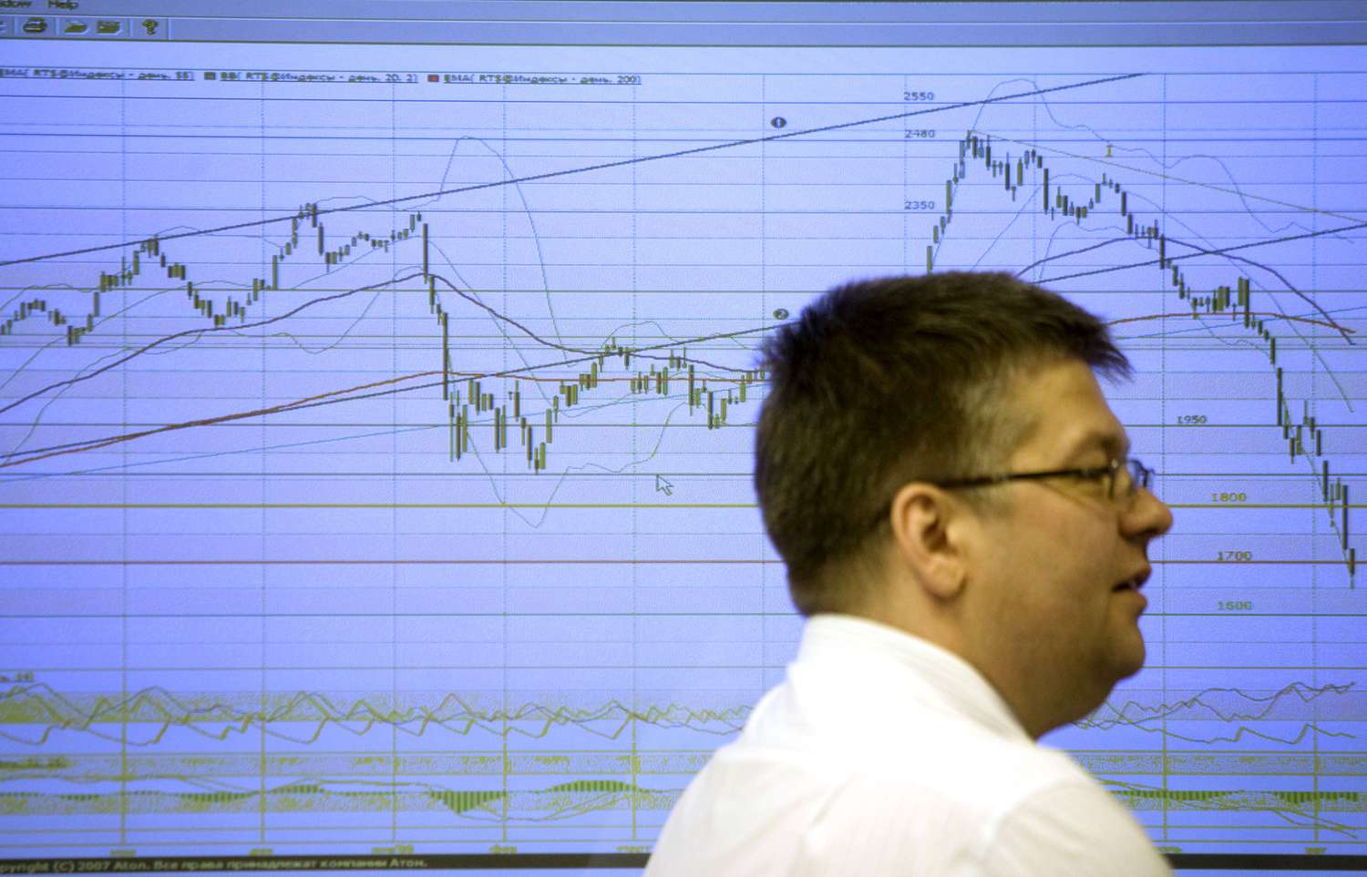 A news anchor sits in front of a screen showing a large graph of a stock's trading range data.