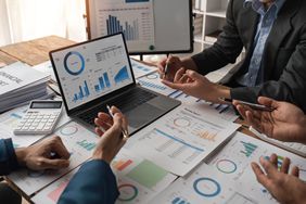 A group of financial analysts meet around a table cluttered with financial documents to discuss the merits of various exchange-listed securities that they can’t trade on a centralized exchange.