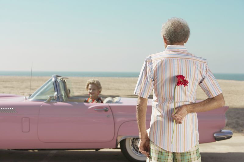 An older person, flower behind their back, looking at a date seated in a pink cadillac.