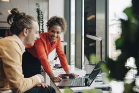 A man and a woman in an office laugh together over something they have seen on a laptop screen.