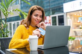 Young woman uses credit card while looking at her laptop at a table outside, with a thoughtful look on her face.