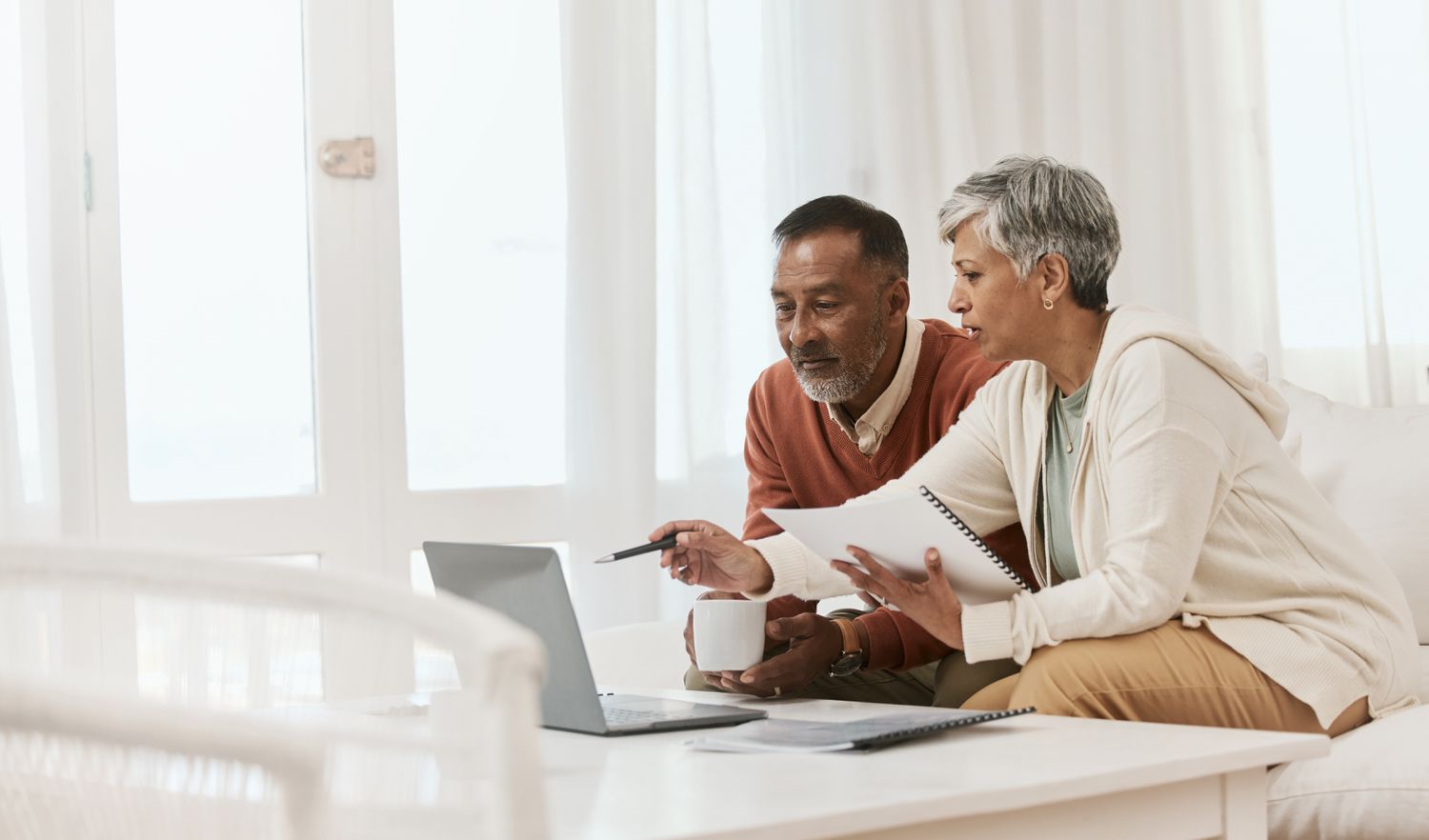 A mature couple reviews finances on a laptop while sitting in a modern living room.