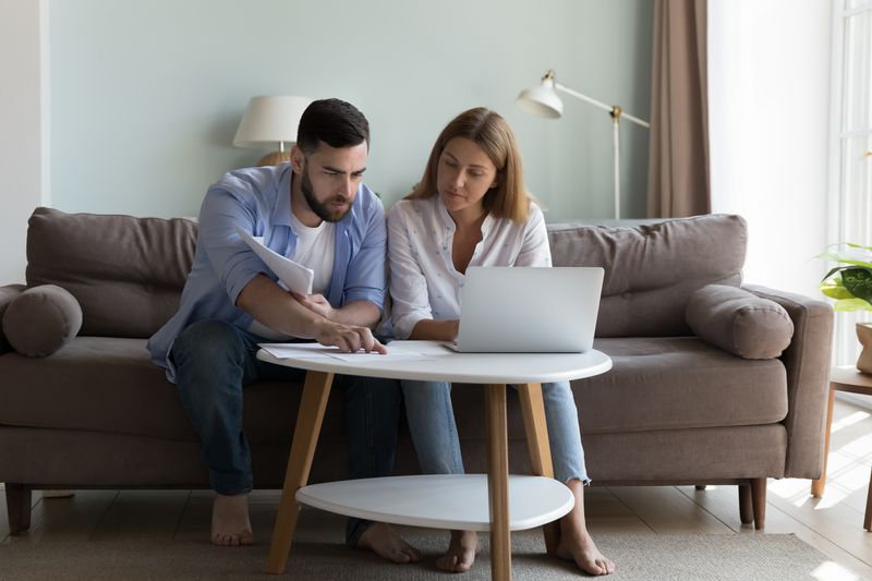 Millennial couple sitting on living room sofa and studying laptop and paperwork together