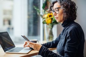 Mature woman sitting at cafe looking at her cell phone while working on laptop computer.