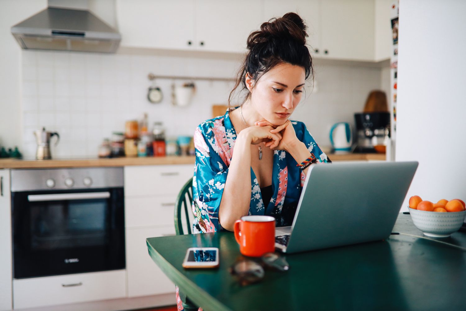 A woman in a blue and pink flowered robe sits at a kitchen table looking at a laptop. There is a phone, a red mug, and a bowl or oranges next to her.