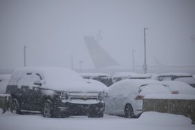 Snow covered cars sit in an airport parking lot, with the tail of a plane visible in the background.