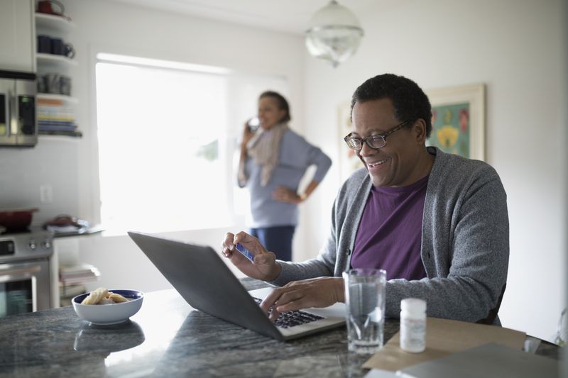 A smiling man with credit card paying bills online at laptop at kitchen counter.
