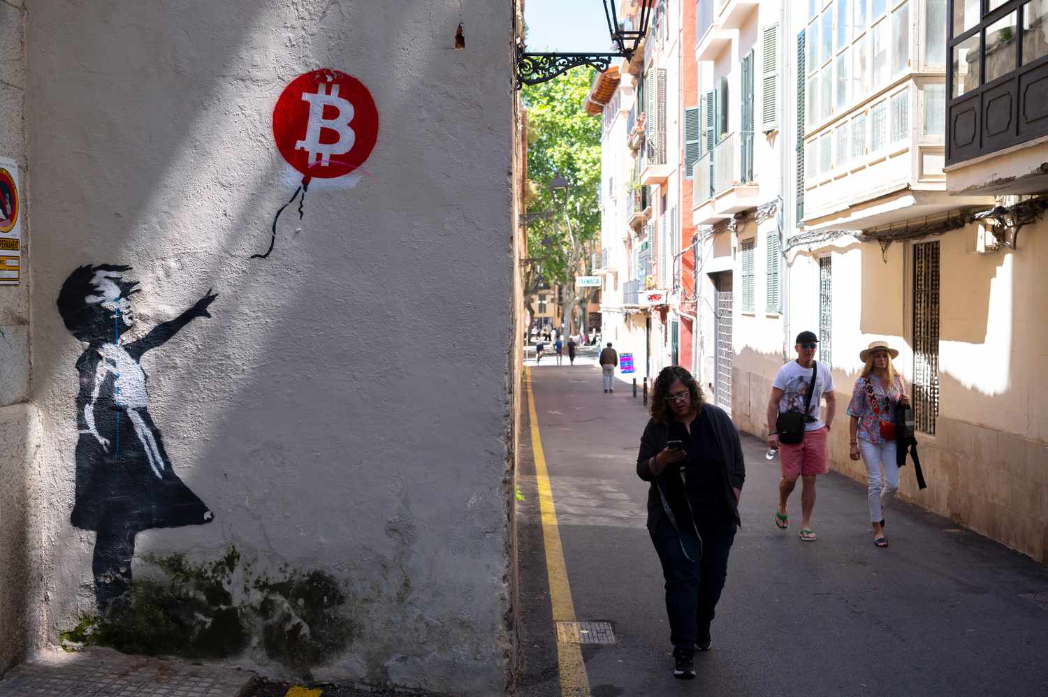 People walk past a Banksy-like depiction on wall near an alley of a young girl letting go of a balloon with a Bitcoin symbol on it.