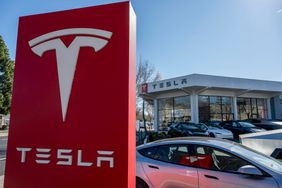The Tesla logo is seen on a red sign with Tesla vehicles in front of a dealership building.