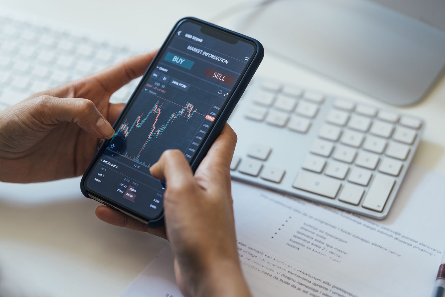 A person sitting at a computer and using a cell phone to place a sell order on stock