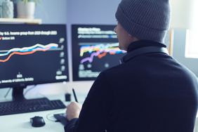 Man wearing a wool hat, sitting with his back to the camera looking at two trading screens. 
