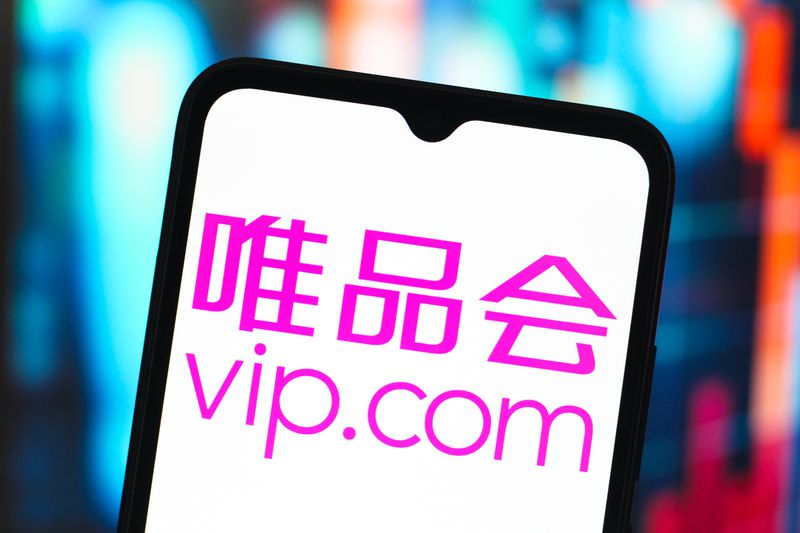 The pink Vipshop Holdings logo is seen on a phone screen in front of a blue and orange background.