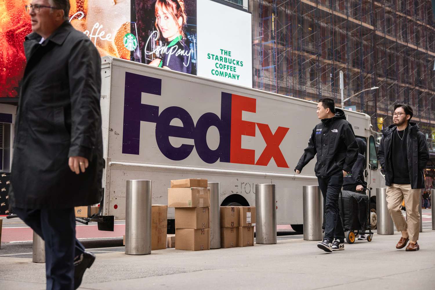 People walk by a stack of packages next to a white FedEx delivery truck on the street.