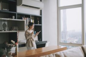 A woman stands in her apartment sipping from a mug and looking at her phone