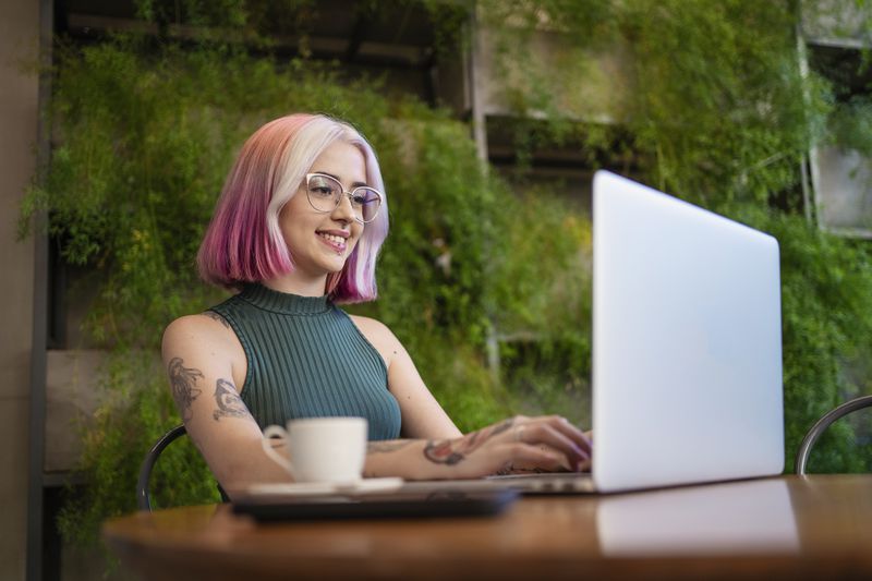 A woman with pink hair and tattoos works on a computer with a coffee cup next to her.