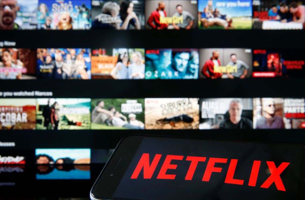 Close-up of iPhone displaying Netflix logo, with Netflix app displayed on a TV monitor in the background. 