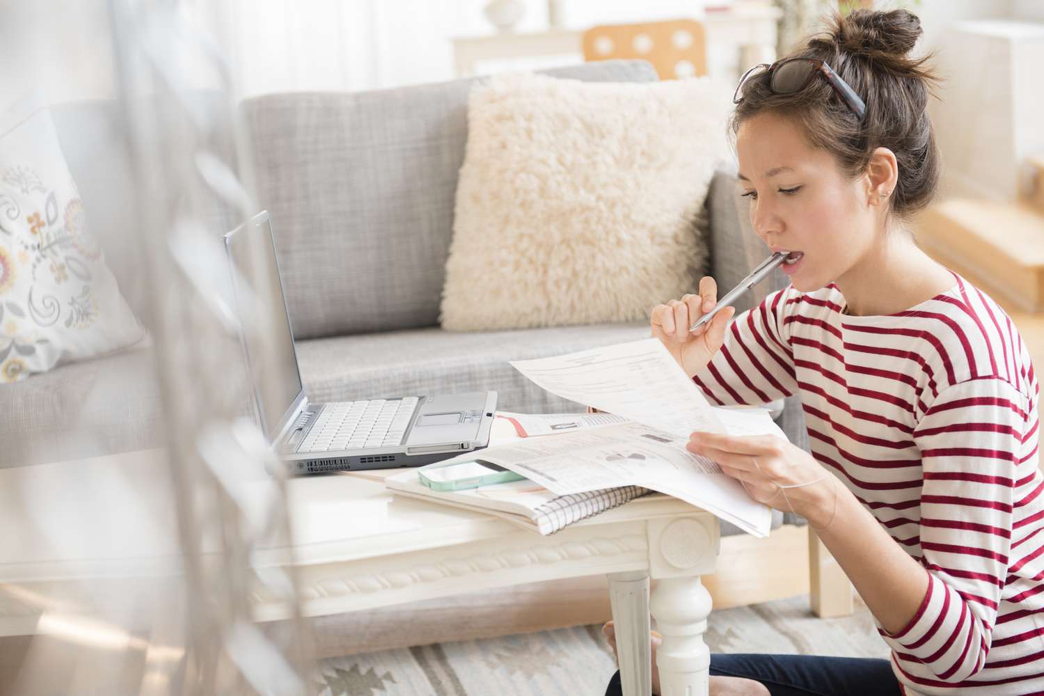 A young woman with her hair tied up and sunglasses perched on her head sits on the floor near a low table in a living room. She is wearing a red and white striped shirt and holding a pen to her mouth while concentrating on papers in her hands. A laptop, notebook, and other documents are spread out on the table in front of her, suggesting she is working or studying. The cozy room features a gray couch with decorative pillows and a fluffy beige cushion in the background.