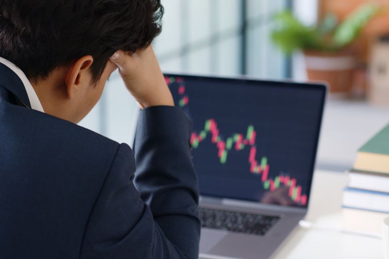 A man wearing a suit is pictured from behind with his head in his hands as he looks at a computer screen displaying declining stock prices.
