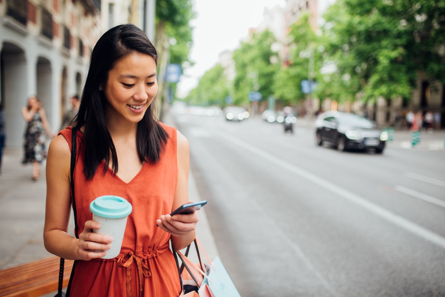 A woman in an orange dress stands on a street with a coffee and smartphone in hand