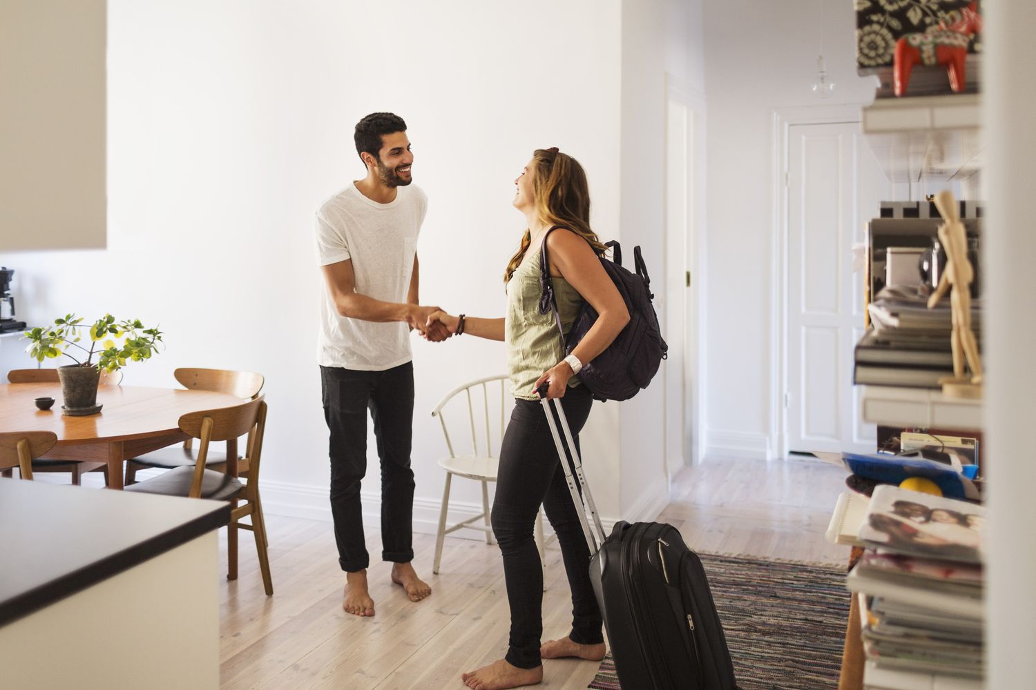 A woman with rolling luggage greets a man in a white t-shirt in an apartment.