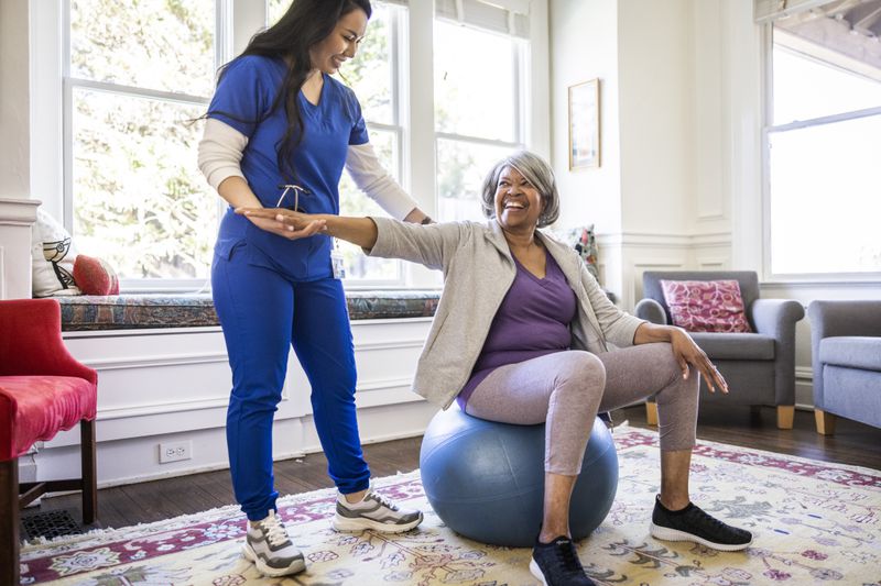 Gray-haired woman sits on a therapy ball while a physical therapist holds her arm up