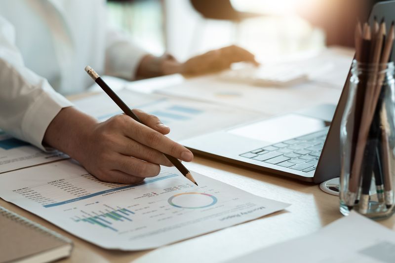 Close up of hand holding a pencil over paper showing financial data next to a laptop