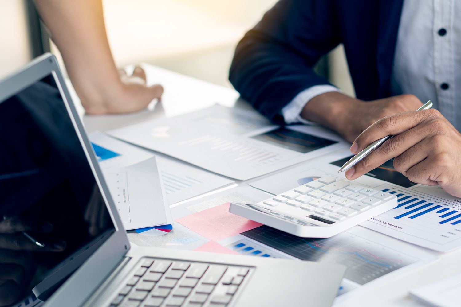 Two business professionals work at a desk covered with paperwork, a computer, and a calculator to finalize the company semiannual report to stockholders.