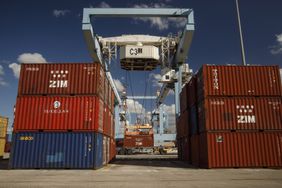 Shipping containers stacked near a port crane under a clear sky at a shipping dock