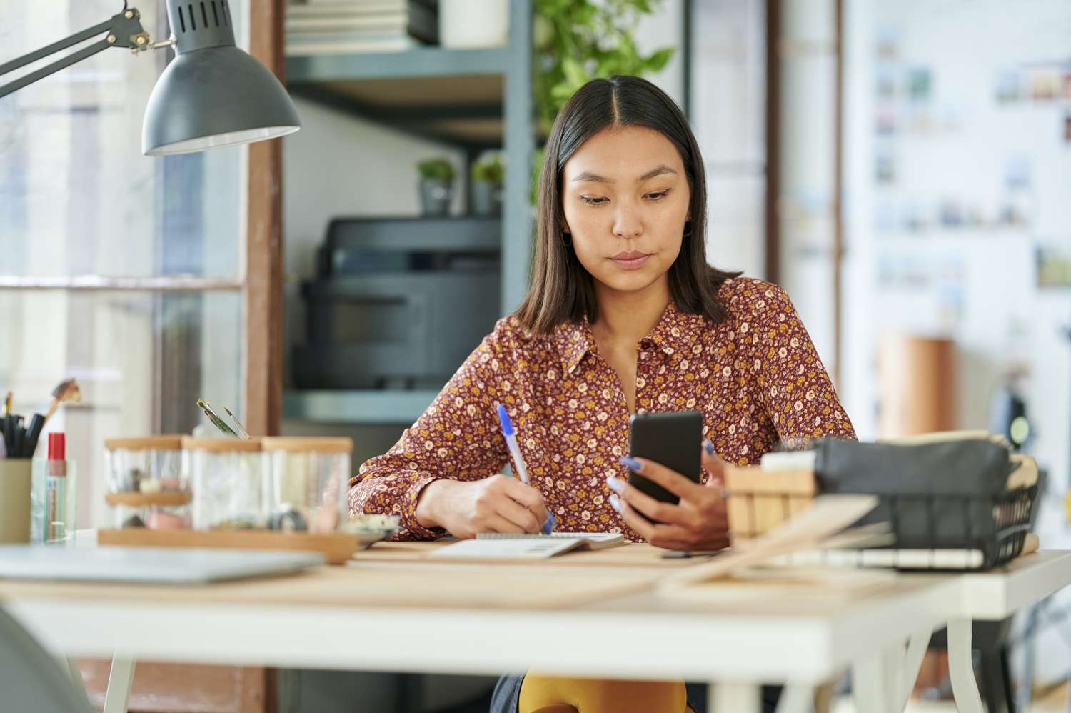 Young woman sitting at her home desk and looking seriously at her phone, with pen and paper in hand
