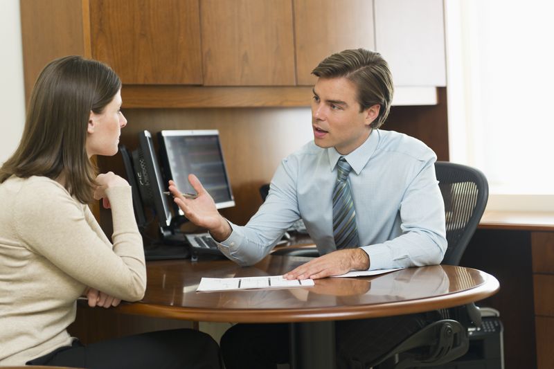 A salesperson at an office desk motions with their hand as they attempt to coerce a reluctant customer into buying an additional product from their company.