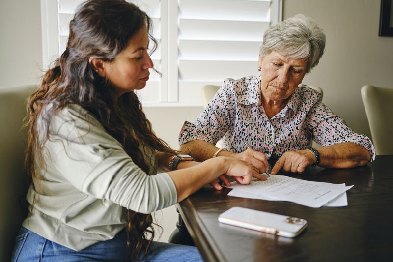 A senior woman signing paperwork with another person present.