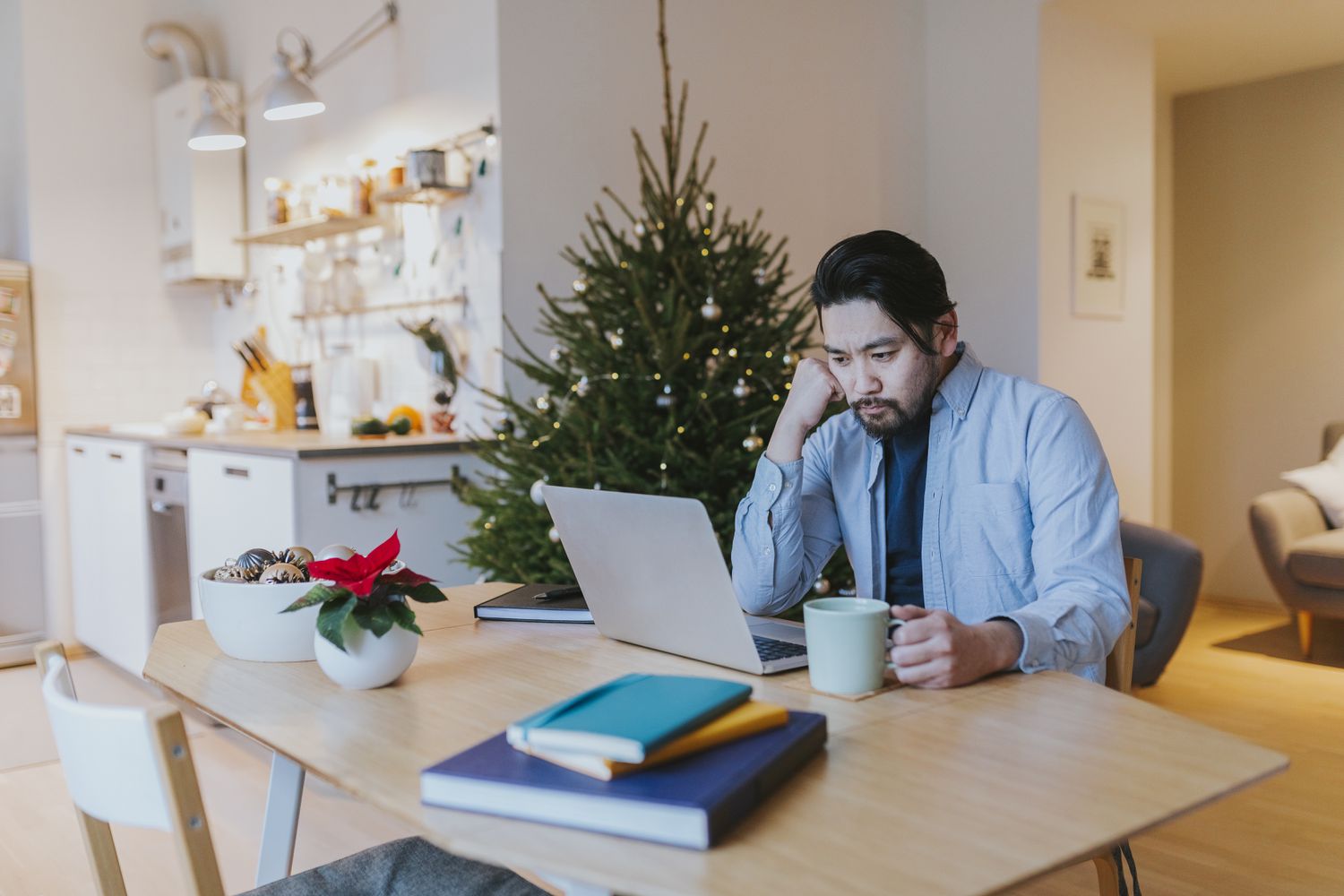 Young Asian man with coffee at dining room table, looking intently at his laptop screen. 