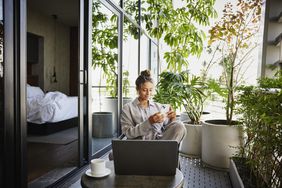 Person in pajamas seated on a balcony using a phone, surrounded by plants, and a laptop on a small table