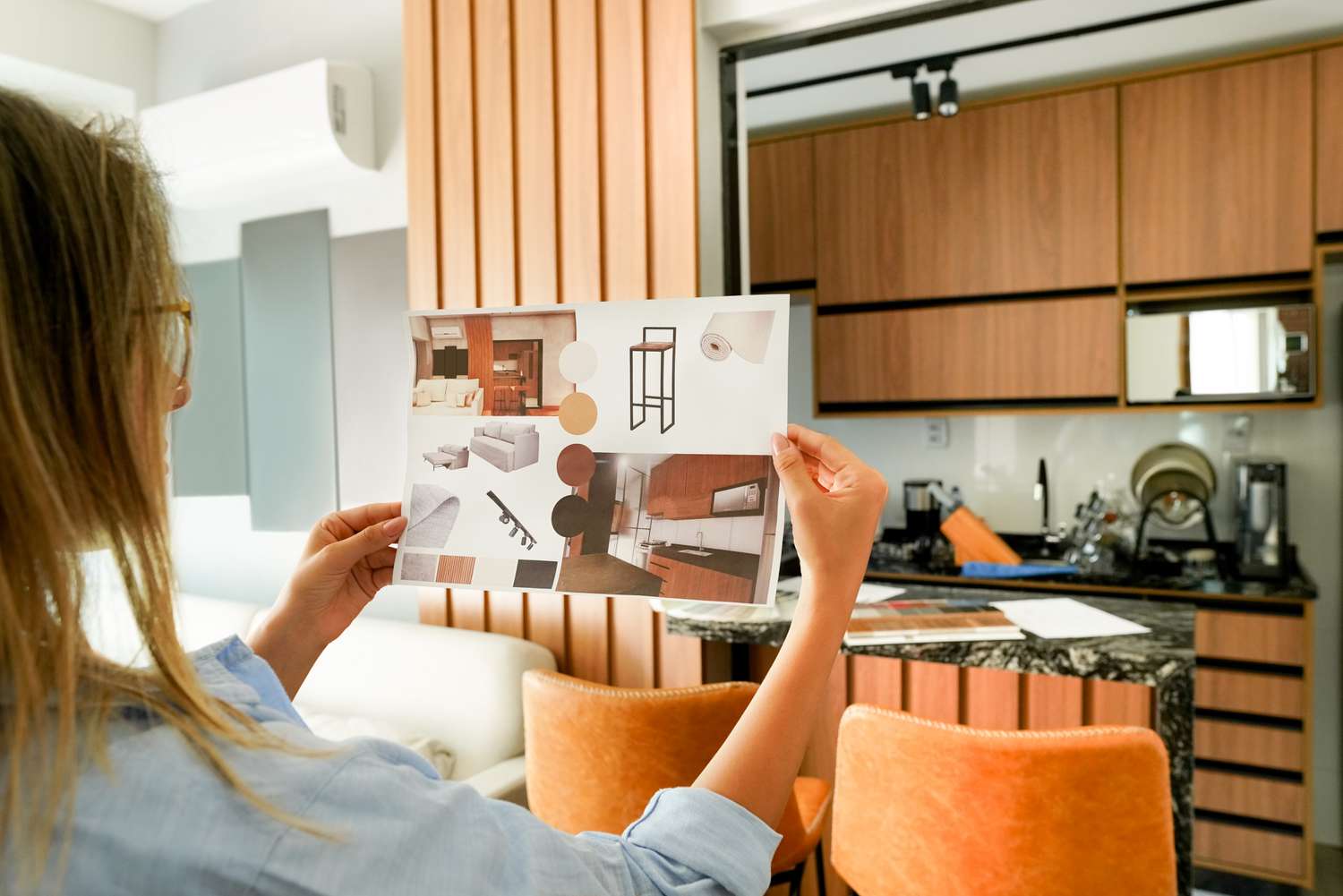 A woman holding an interior design mood board in a modern kitchen, with a focus on wood and neutral tones. The mood board features images and swatches of furniture, fabrics, and colors that complement the kitchen’s design.