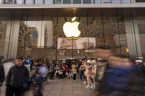 Crowds of people pass by the glass exterior of an Apple store, displaying the company's logo.