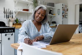 A woman at a laptop in a kitchen taking notes on paper