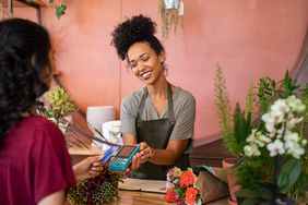 Small business owner requests payment from customer by holding card reader machine while customer uses card. They are both in a flower shop.