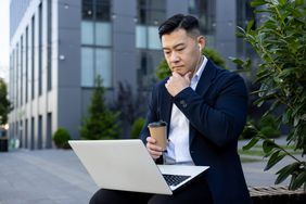 Man sitting on a bench outside an office building, looking at a laptop.